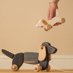 Hand holding a Bajo wooden dachshund puppy toy above a Bajo pull along dachshund on a wooden floor