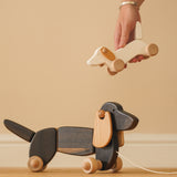Hand holding a Bajo wooden dachshund puppy toy above a Bajo pull along dachshund on a wooden floor