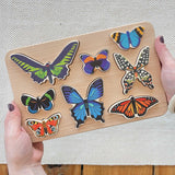 The Bajo World Of Butterflies Sorter Puzzle shown in an adult's hand. This puzzle has butterfly shaped pieces that are decorated on both sides that slot into the coordinating shapes on the base. These heirloom quality toys by Bajo are part of a wide range of wooden toys available here at Babipur.