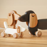 A Bajo white wooden dachshund puppy standing next to the dark oak and natural dachshund pull along dogs available at Babipur.