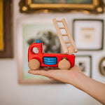 Hand holding Bajo sustainable wooden fire engine toy in front of photo frames in the background