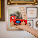 Hand holding Bajo sustainable wooden fire engine toy in front of photo frames in the background