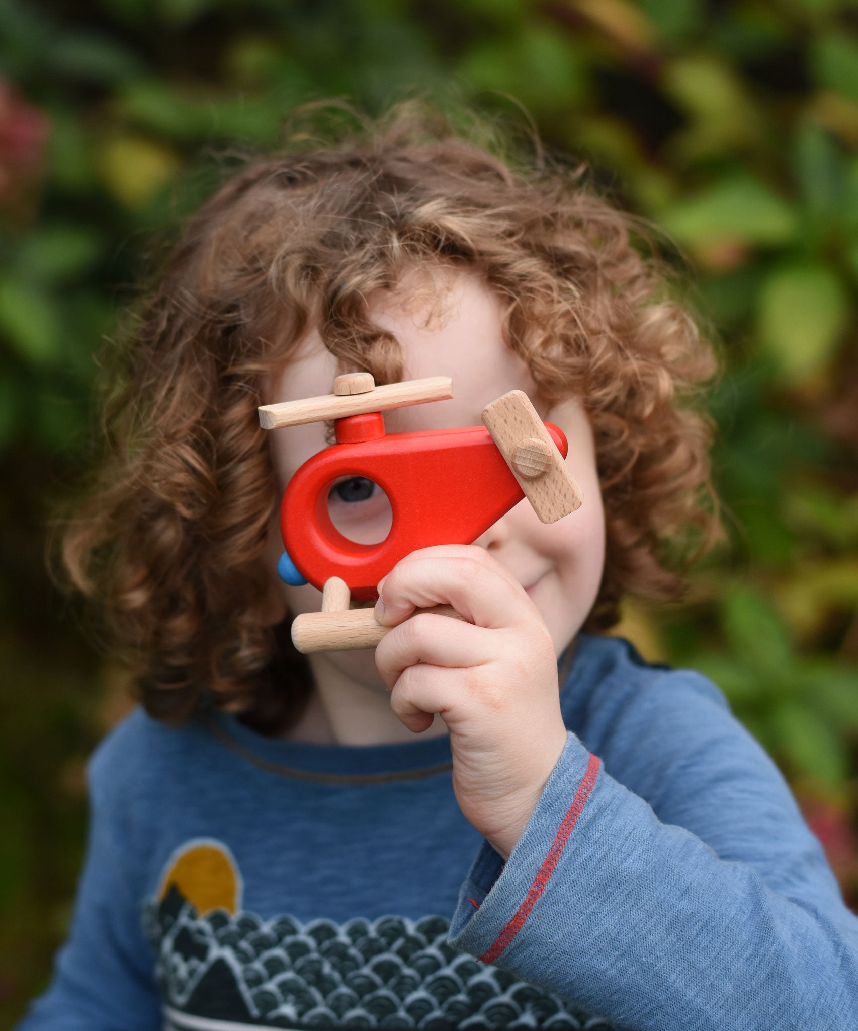 A child looking through the circle shaped hole in the Bajo Red Fire Engine Helicopter wooden toy. The red coloured wooden helicopter has natural propellers and stand and a blue lamp details on the front. These heirloom quality toys by Bajo are part of a wide range of wooden toys available here at Babipur. 