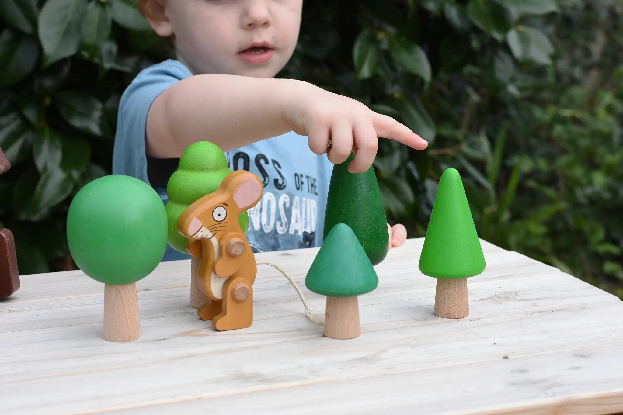 Child playing with the Bajo green forest tree set - 5 wooden trees in a set of 5 placed on a wooden surface with the Gruffalo mouse character figure. 
