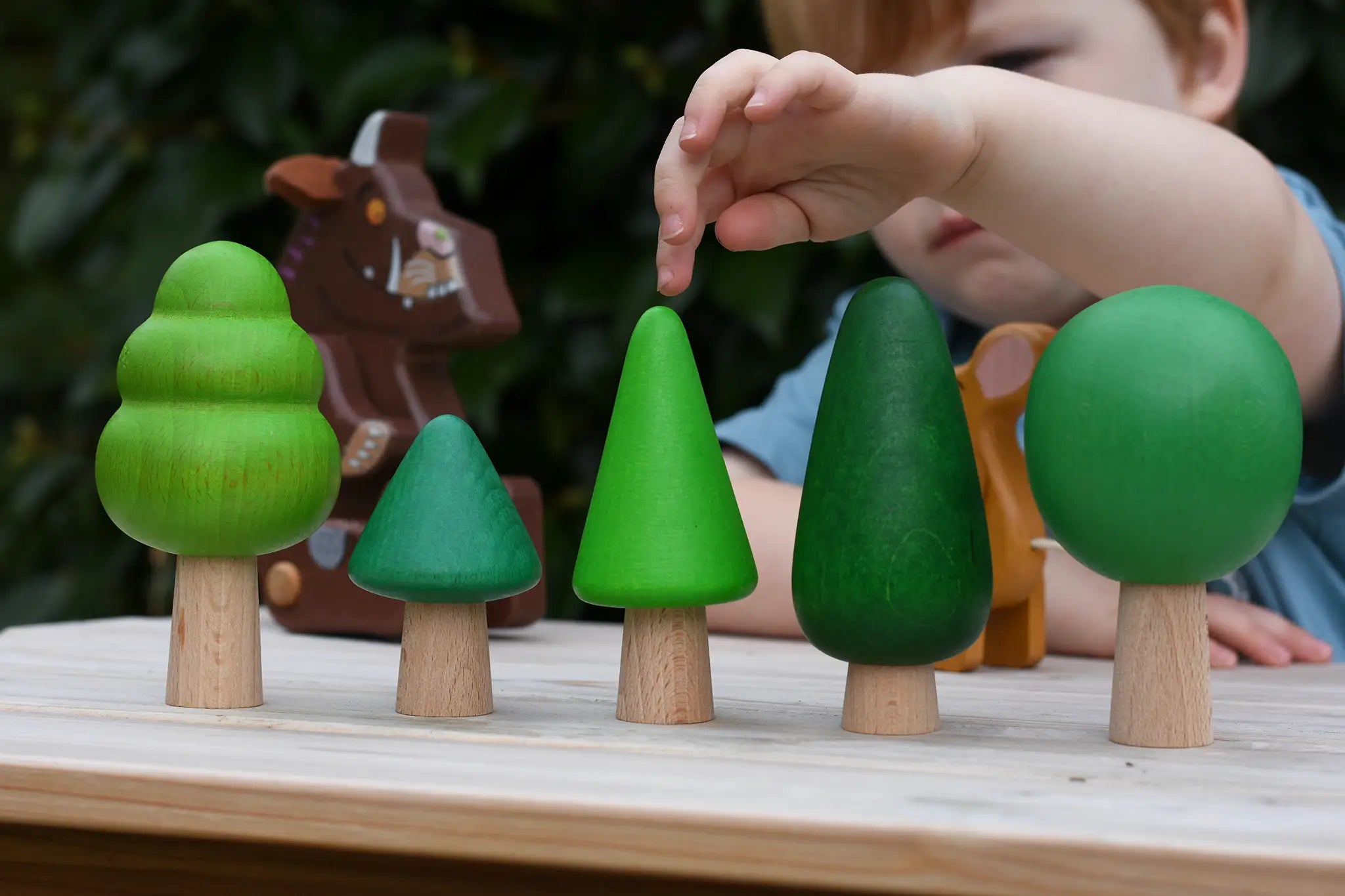 Child playing with the Bajo green forest tree set lining them up on a table. Gruffalo figure in the background.