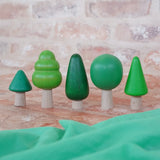 Bajo forest wooden toy tree figure set. Five green trees in different heights and shapes. Green laura's play cloth can be seen in the foreground.