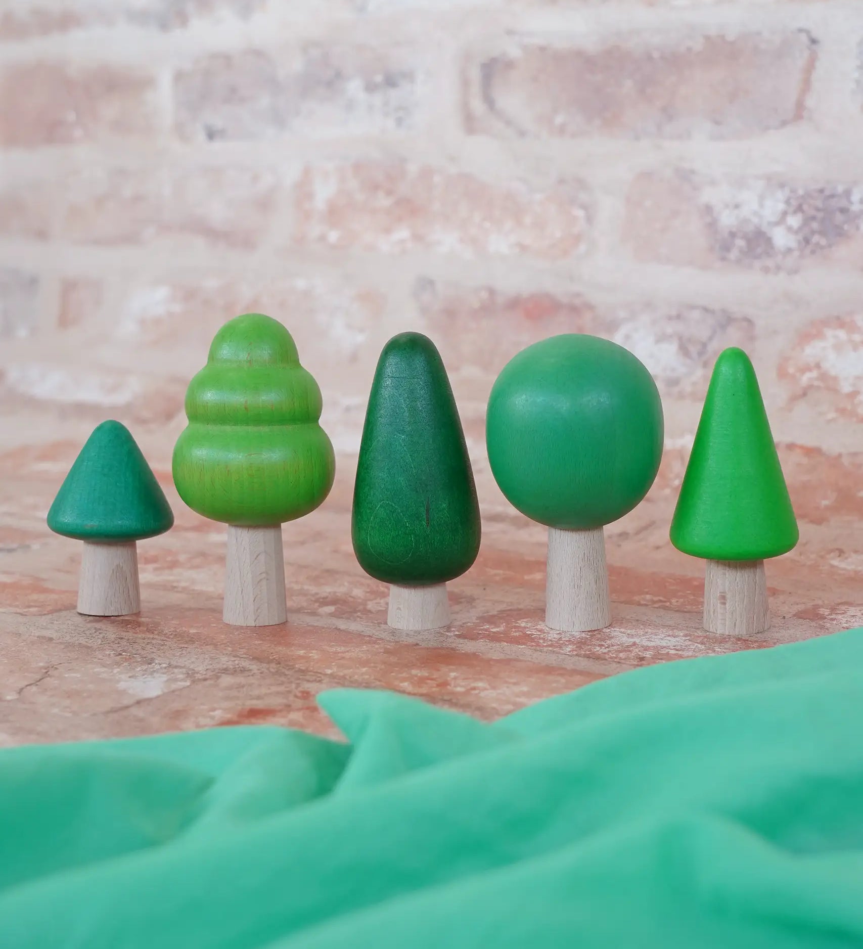 Bajo forest wooden toy tree figure set. Five green trees in different heights and shapes. Green laura's play cloth can be seen in the foreground.