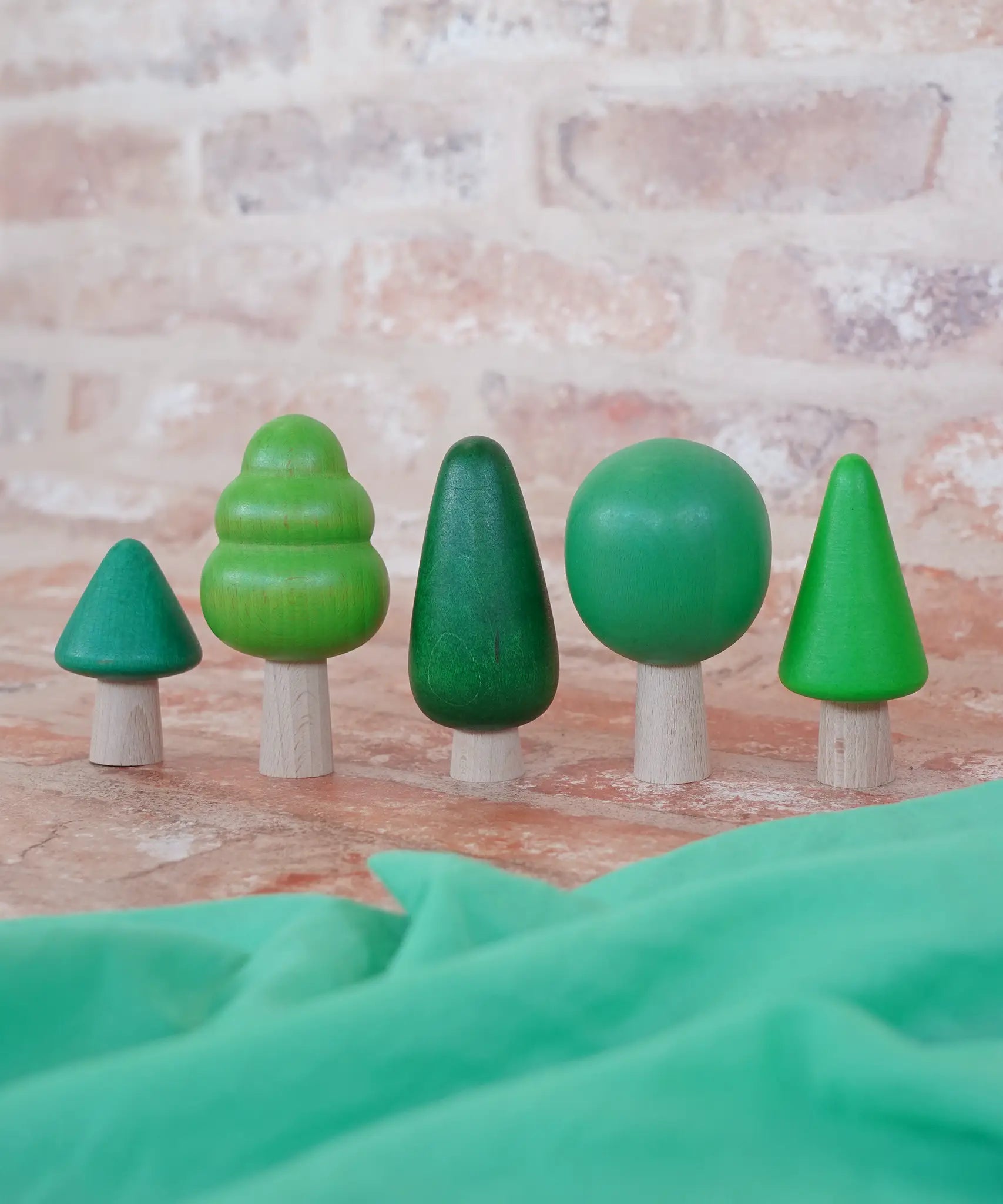 Bajo forest wooden toy tree figure set. Five green trees in different heights and shapes. Green laura's play cloth can be seen in the foreground.