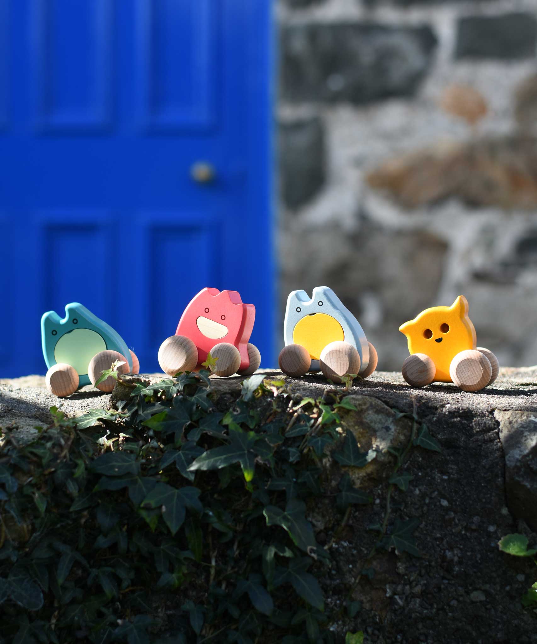 Four Bajo push along wooden ghost toys paced on a stone wall outdoors. Toe toy has a yellow painted body and natural wooden wheels. These heirloom quality toys by Bajo are part of a wide range of wooden toys available here at Babipur. 