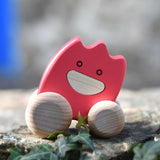 A pink coloured Bajo wooden ghost toy placed on a stone wall outdoors.  The toy has a pink painted body and natural wooden wheels. These heirloom quality toys by Bajo are part of a wide range of wooden toys available here at Babipur. 