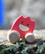 A pink coloured Bajo wooden ghost toy placed on a stone wall outdoors.  The toy has a pink painted body and natural wooden wheels. These heirloom quality toys by Bajo are part of a wide range of wooden toys available here at Babipur. 