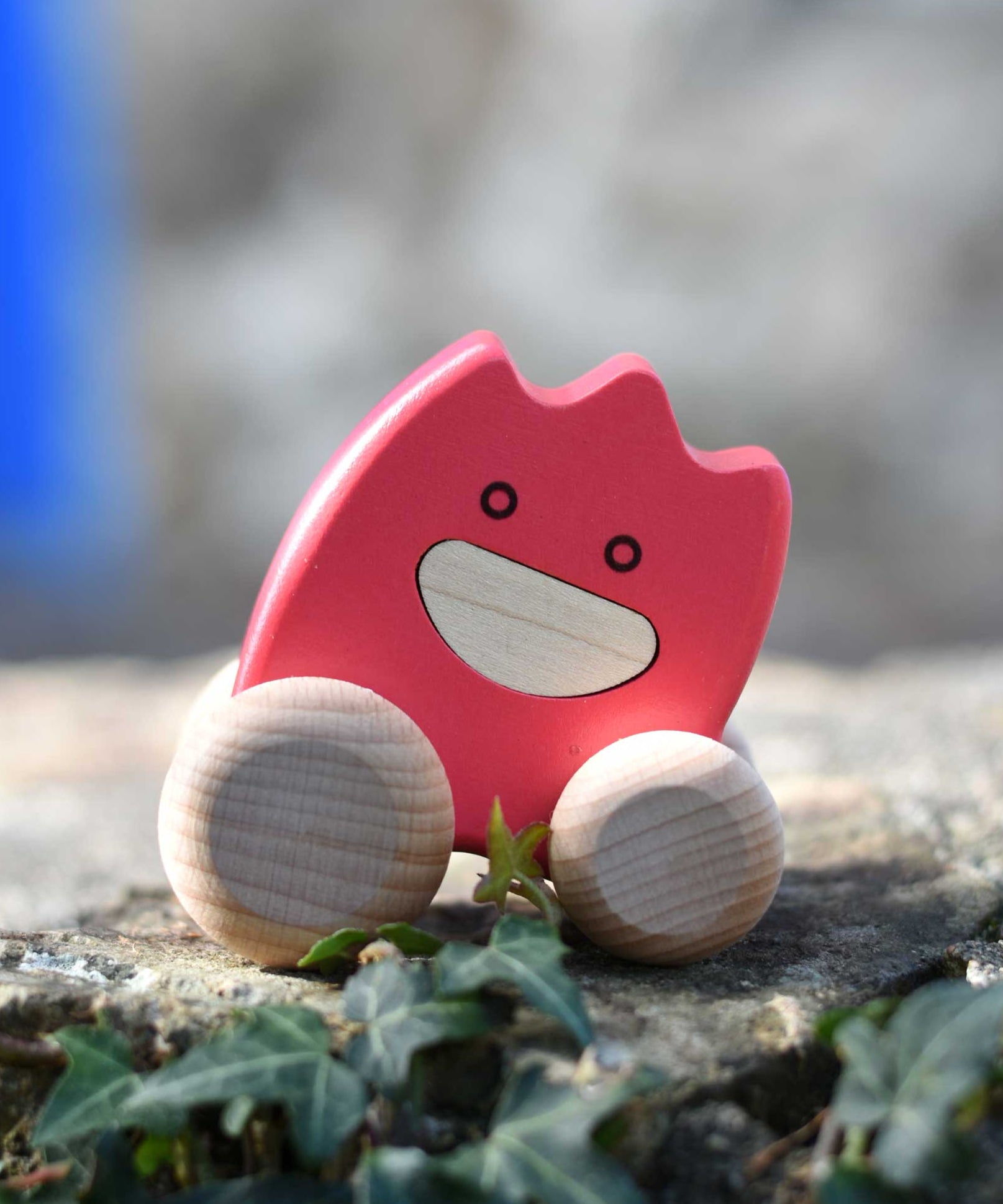 A pink coloured Bajo wooden ghost toy placed on a stone wall outdoors.  The toy has a pink painted body and natural wooden wheels. These heirloom quality toys by Bajo are part of a wide range of wooden toys available here at Babipur. 