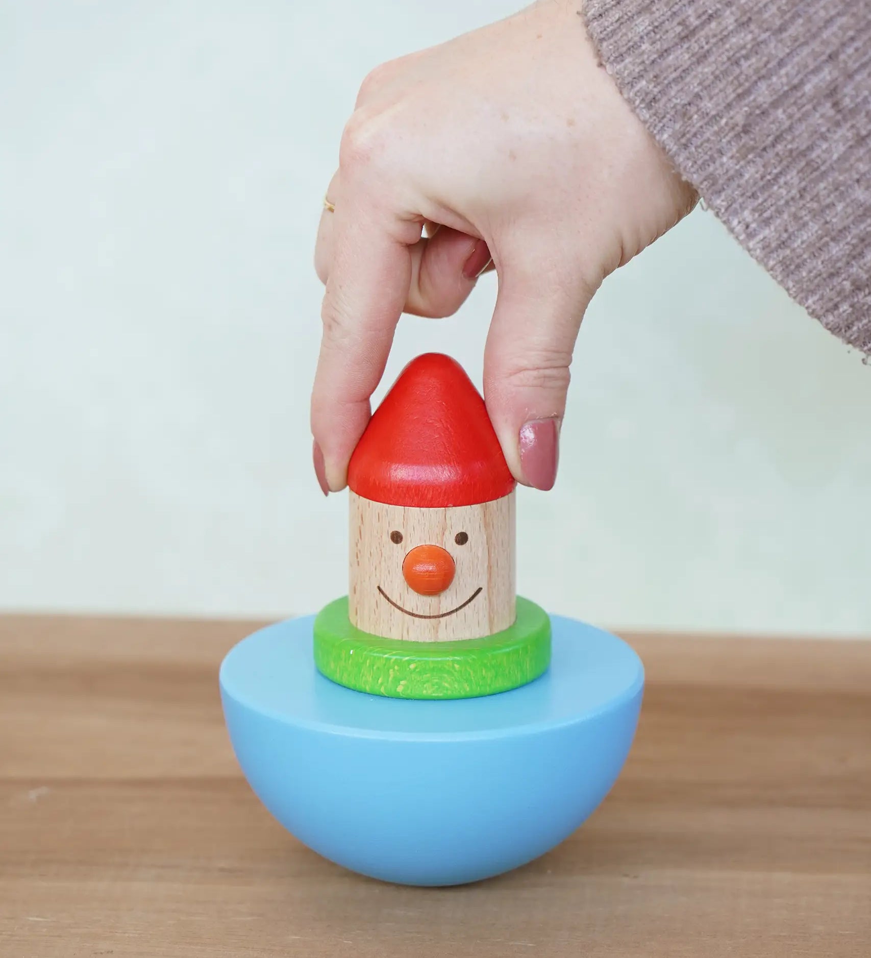 Adult's hand holding the Bajo roly poly wobbling dwarf wooden baby toy upright. Blue half sphere base that wobbles when pushed with a wooden dwarf character on top from Babipur