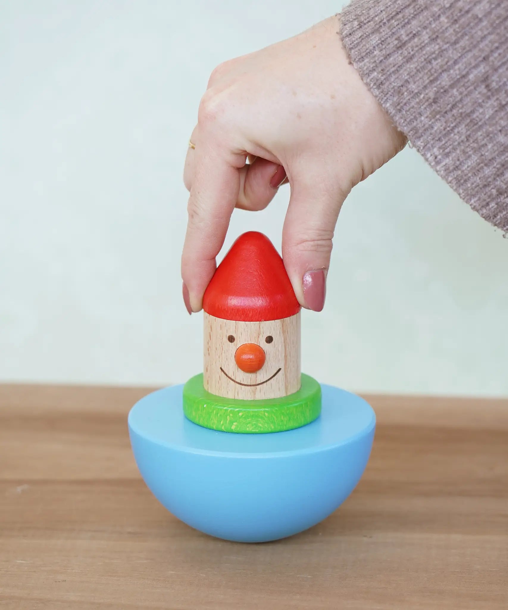 Adult's hand holding the Bajo roly poly wobbling dwarf wooden baby toy upright. Blue half sphere base that wobbles when pushed with a wooden dwarf character on top from Babipur