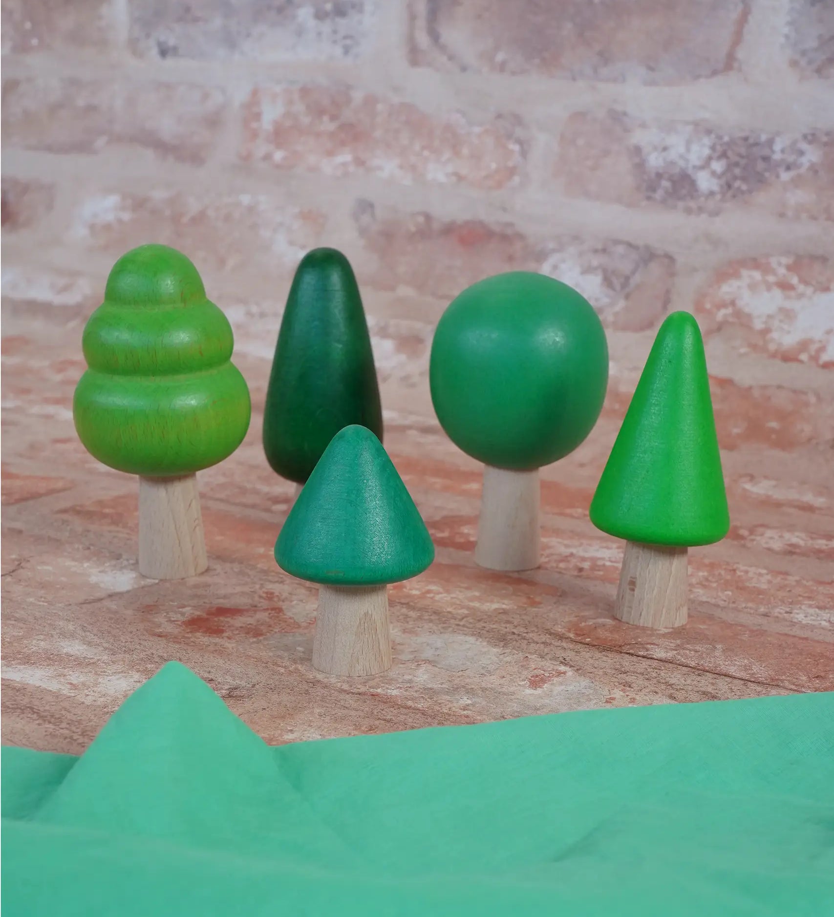 Bajo forest wooden toy tree figure set. Five green trees in different heights and shapes. Green laura's play cloth can be seen in the foreground.