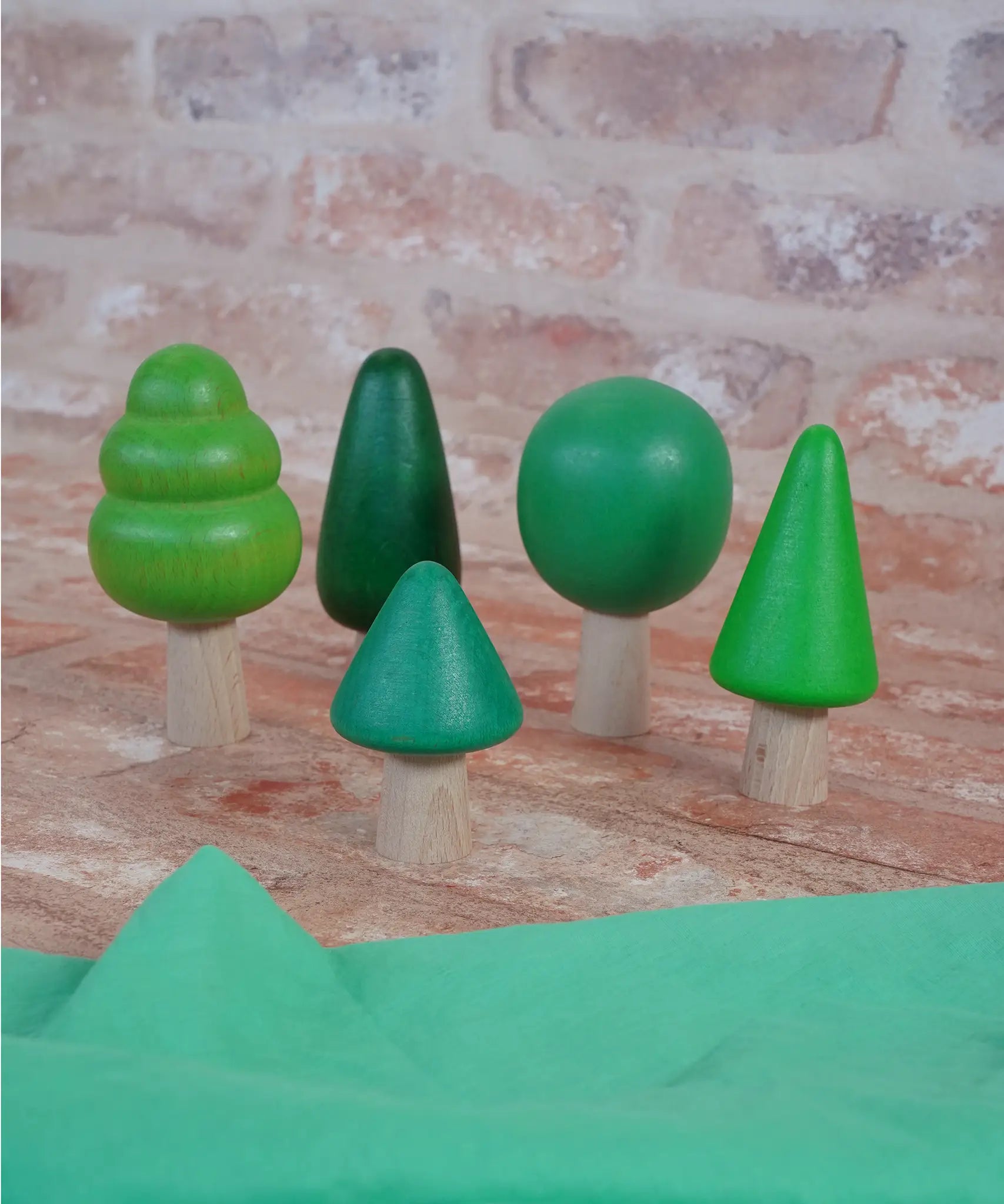 Bajo forest wooden toy tree figure set. Five green trees in different heights and shapes. Green laura's play cloth can be seen in the foreground.