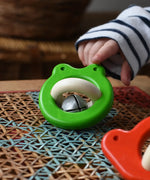 Bajo green Frog Rattle wooden baby toy placed on a rattan stool in the Babipur playroom, a child is pointing at it in the background..