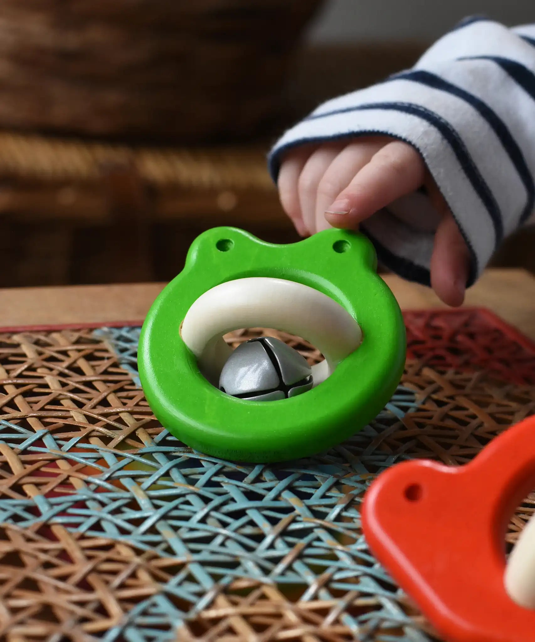 Bajo green Frog Rattle wooden baby toy placed on a rattan stool in the Babipur playroom, a child is pointing at it in the background..