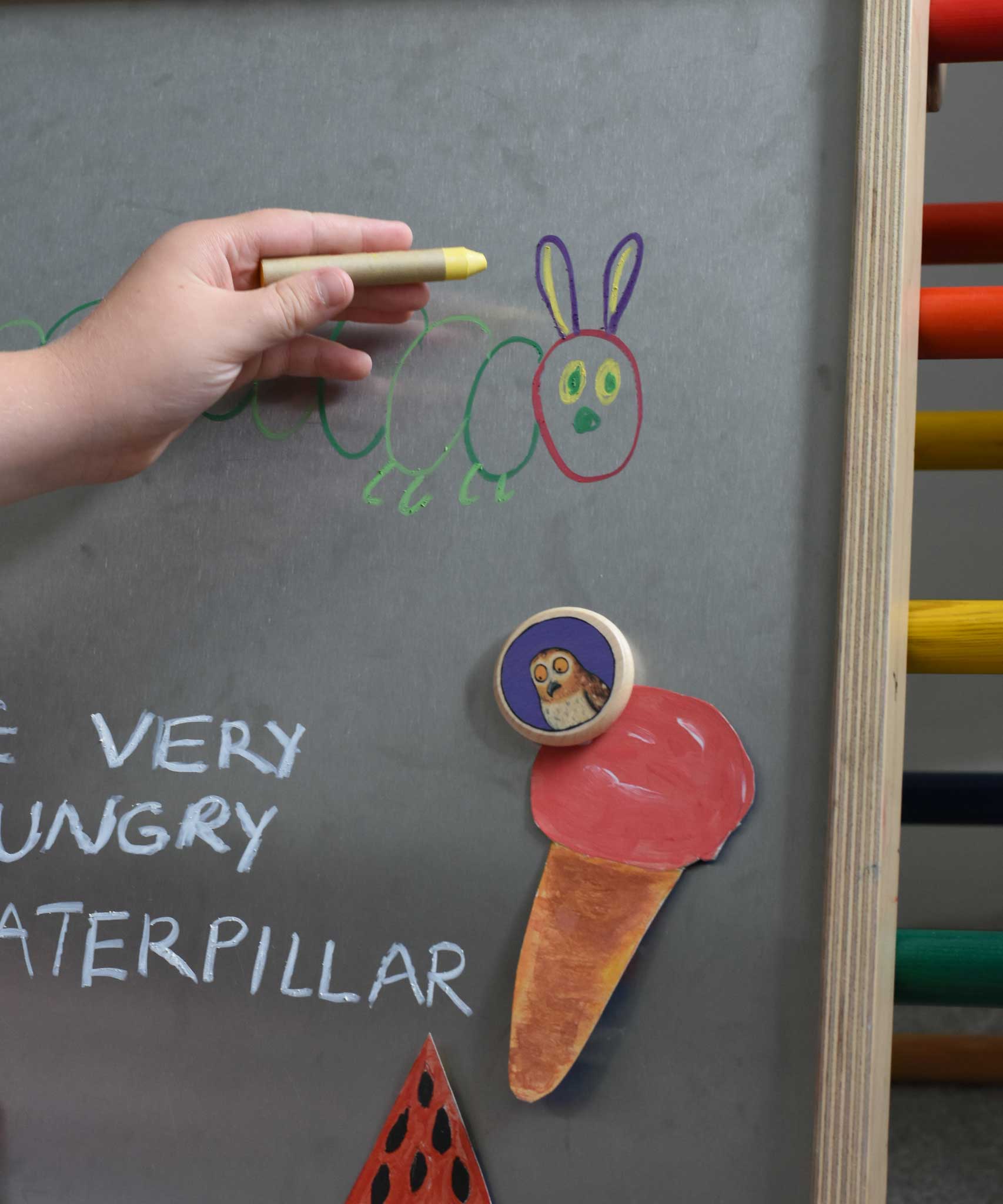A child's hand drawing on a Triclimb biri board. A hand drawnice cream placed on the Triclimb Biri board with an owl Bajo magnet. These magnets are part of a range of Bajo wooden toys available at Babipur. 