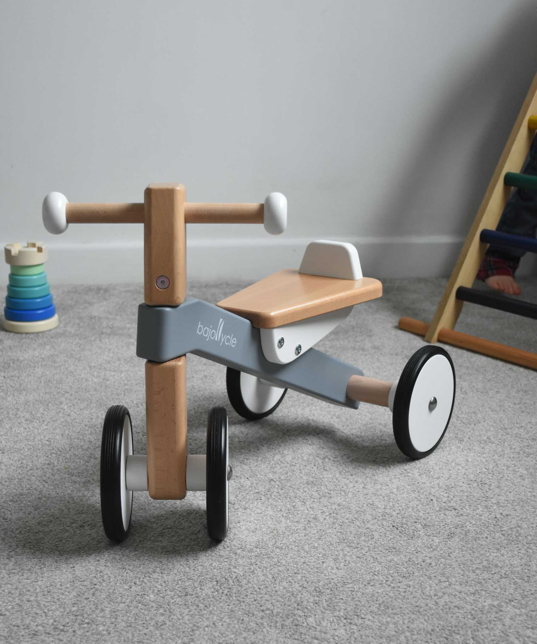 A Bajo Toys wooden Cycle toddler Ride-On in the monochrome, white and grey colourway placed on a carpeted surface. The wooden ride on cycle is part of a wode range of wooden toys available here at Babipur. 