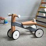 A Bajo Toys wooden Cycle toddler Ride-On in the monochrome, white and grey colourway. The ride on toy has been placed on carpet a child's hand can be seen playing with a wooden stacking toy in the background along with a TriClimb. The wooden ride on cycle is part of a wode range of wooden toys available here at Babipur. 
