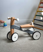 A Bajo Toys wooden Cycle toddler Ride-On in the monochrome, white and grey colourway. The ride on toy has been placed on carpet a child's hand can be seen playing with a wooden stacking toy in the background along with a TriClimb. The wooden ride on cycle is part of a wode range of wooden toys available here at Babipur. 