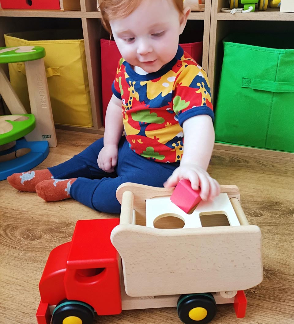 Young boy sat on a wooden floor putting a red toy block into the back of the Bajo red shape sorting toy truck