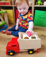 Young boy sat on a wooden floor putting a red toy block into the back of the Bajo red shape sorting toy truck