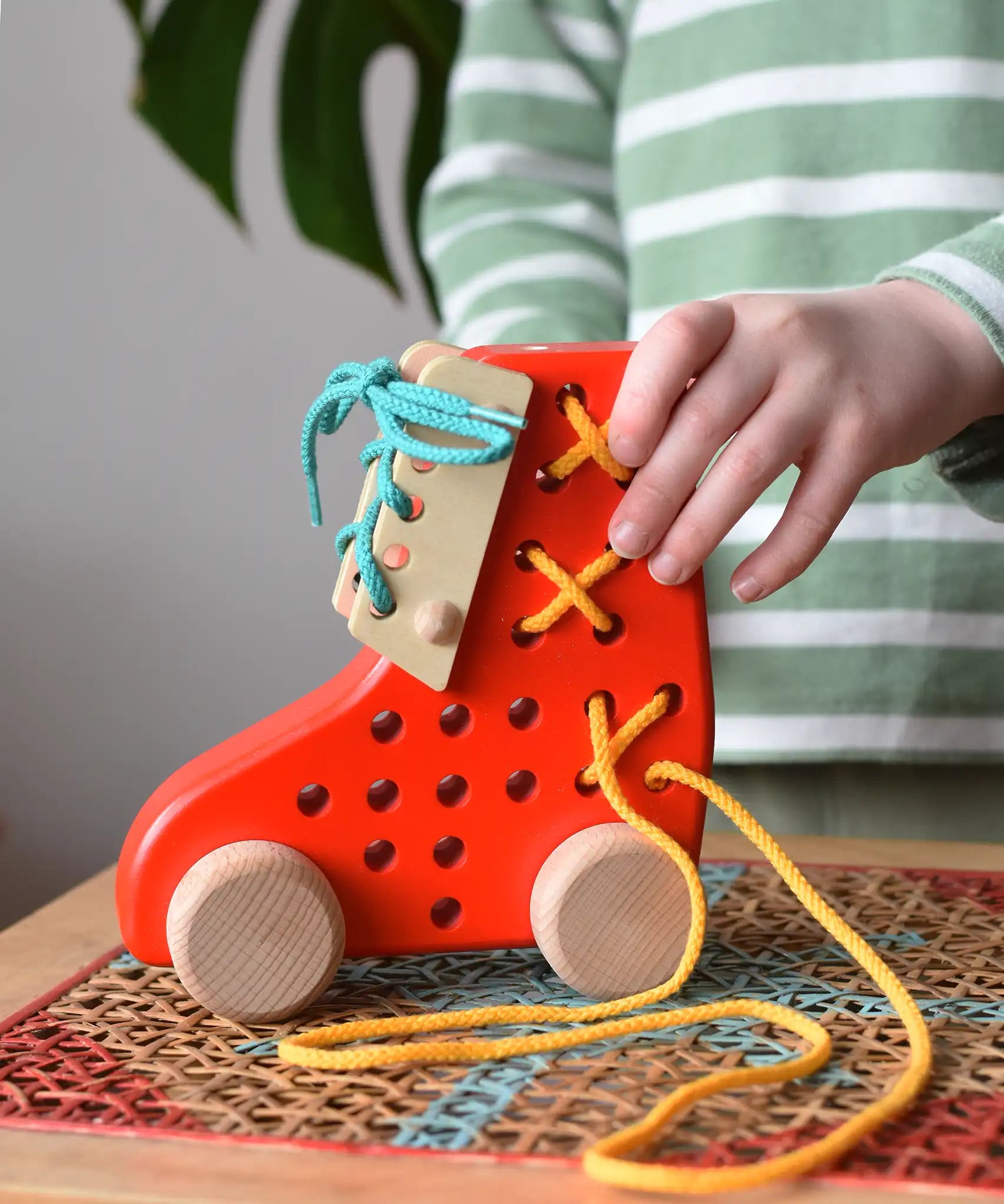 Child playing with the Bajo red lacing shoe wooden push along toy placed on a rattan stool in the Babipur playroom.