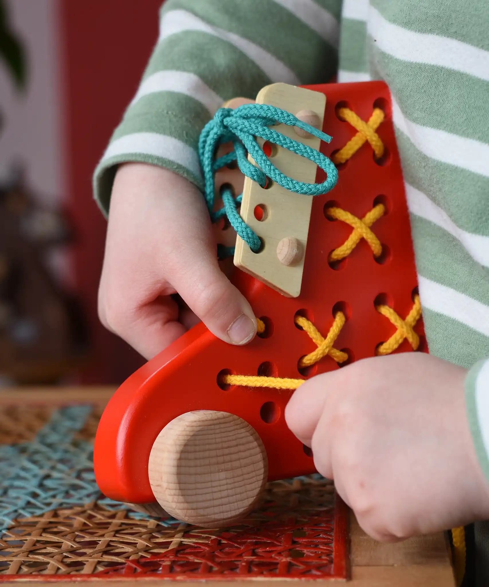 Child playing with the Bajo red lacing shoe wooden push along toy placed on a rattan stool in the Babipur playroom.