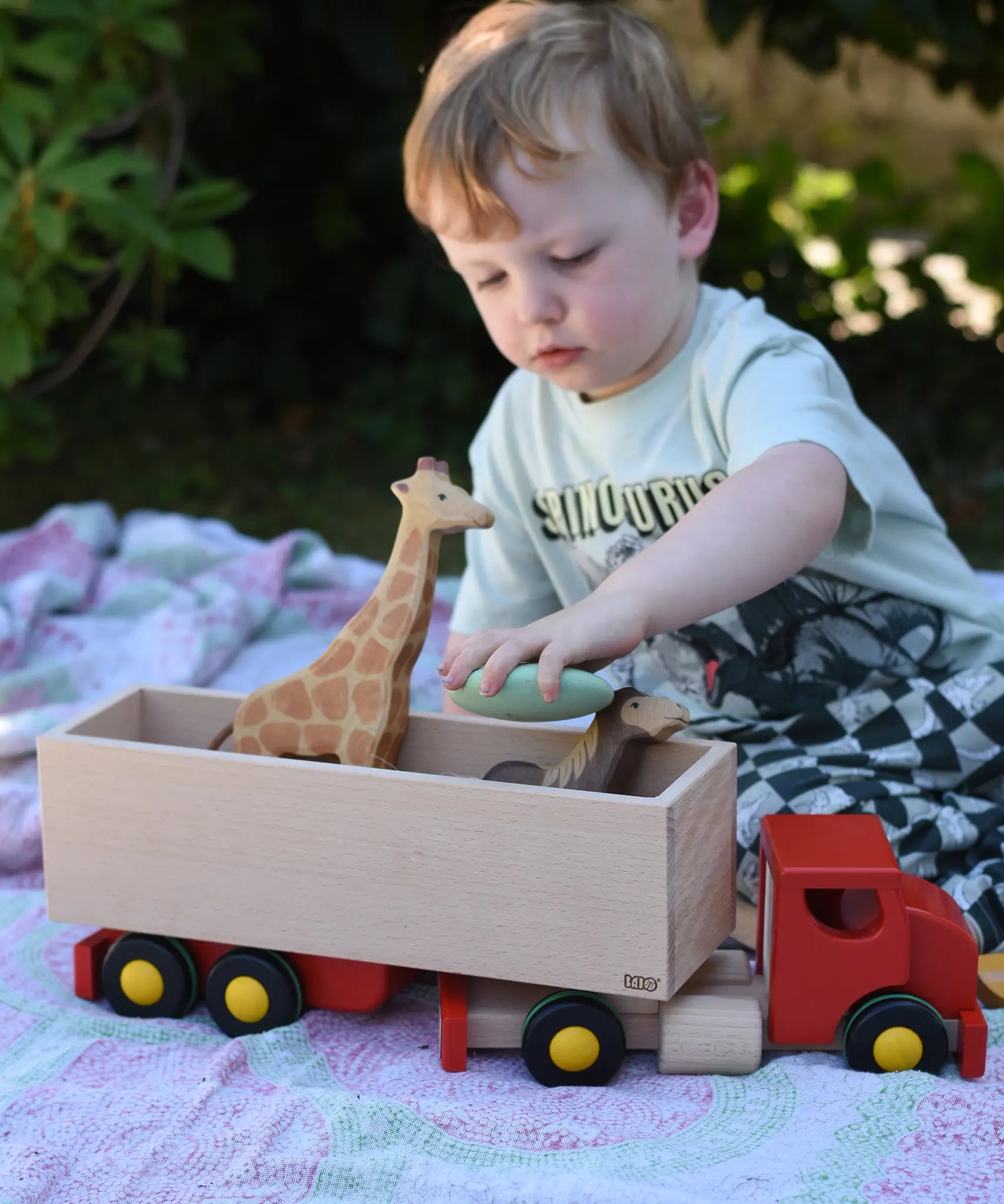 Child playing with the Bajo large truck and trailer wooden toy, placing Bajo pebbles inside with Holztiger animal figures. 