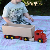 Child playing with the Bajo large truck and trailer wooden toy. Red cab and trailer base and natural wood trailer box.
