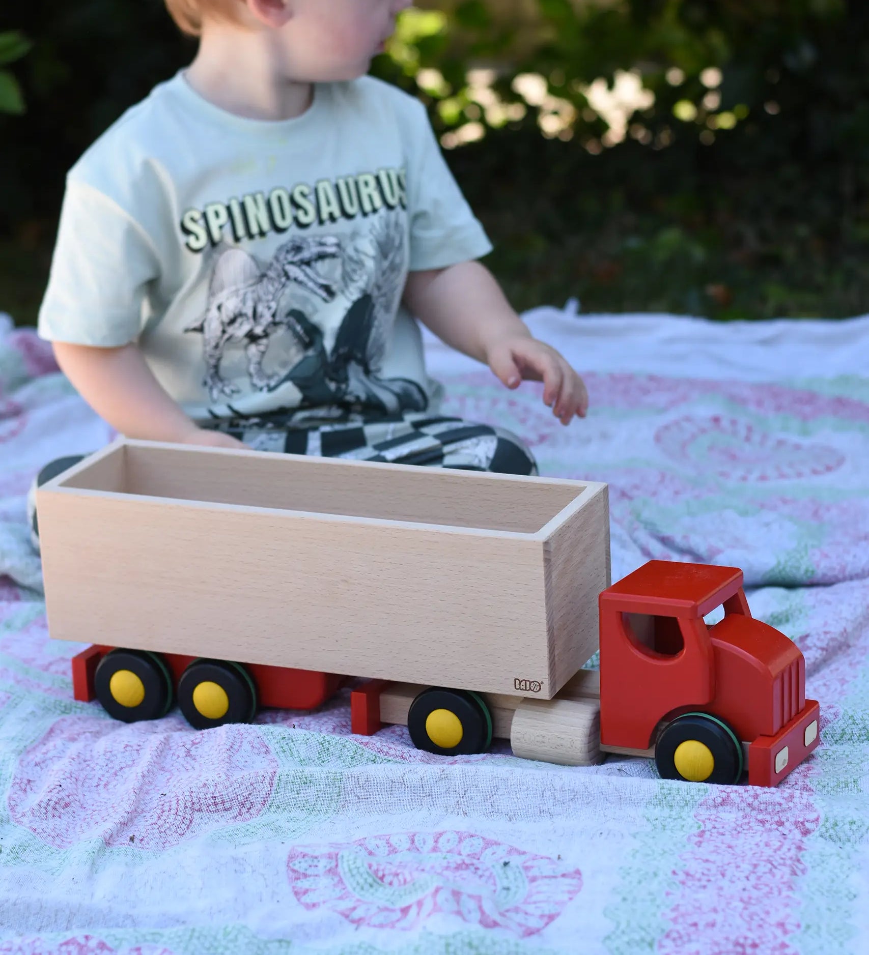 Child playing with the Bajo large truck and trailer wooden toy. Red cab and trailer base and natural wood trailer box.
