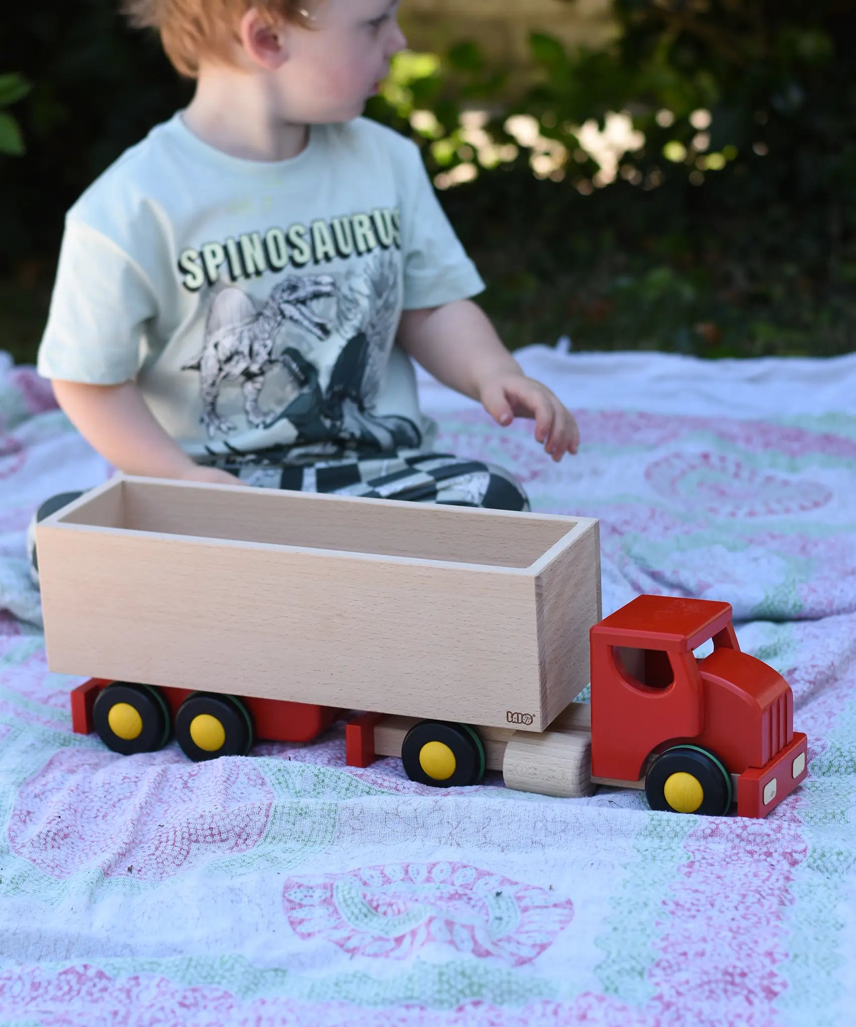 Child playing with the Bajo large truck and trailer wooden toy. Red cab and trailer base and natural wood trailer box.
