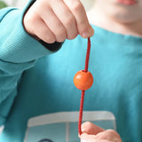 Close up of an orange bead threaded onto a red lace from the Bajo Mathematical Threading Beads Game in a child's hand.  