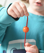 Close up of an orange bead threaded onto a red lace from the Bajo Mathematical Threading Beads Game in a child's hand.  