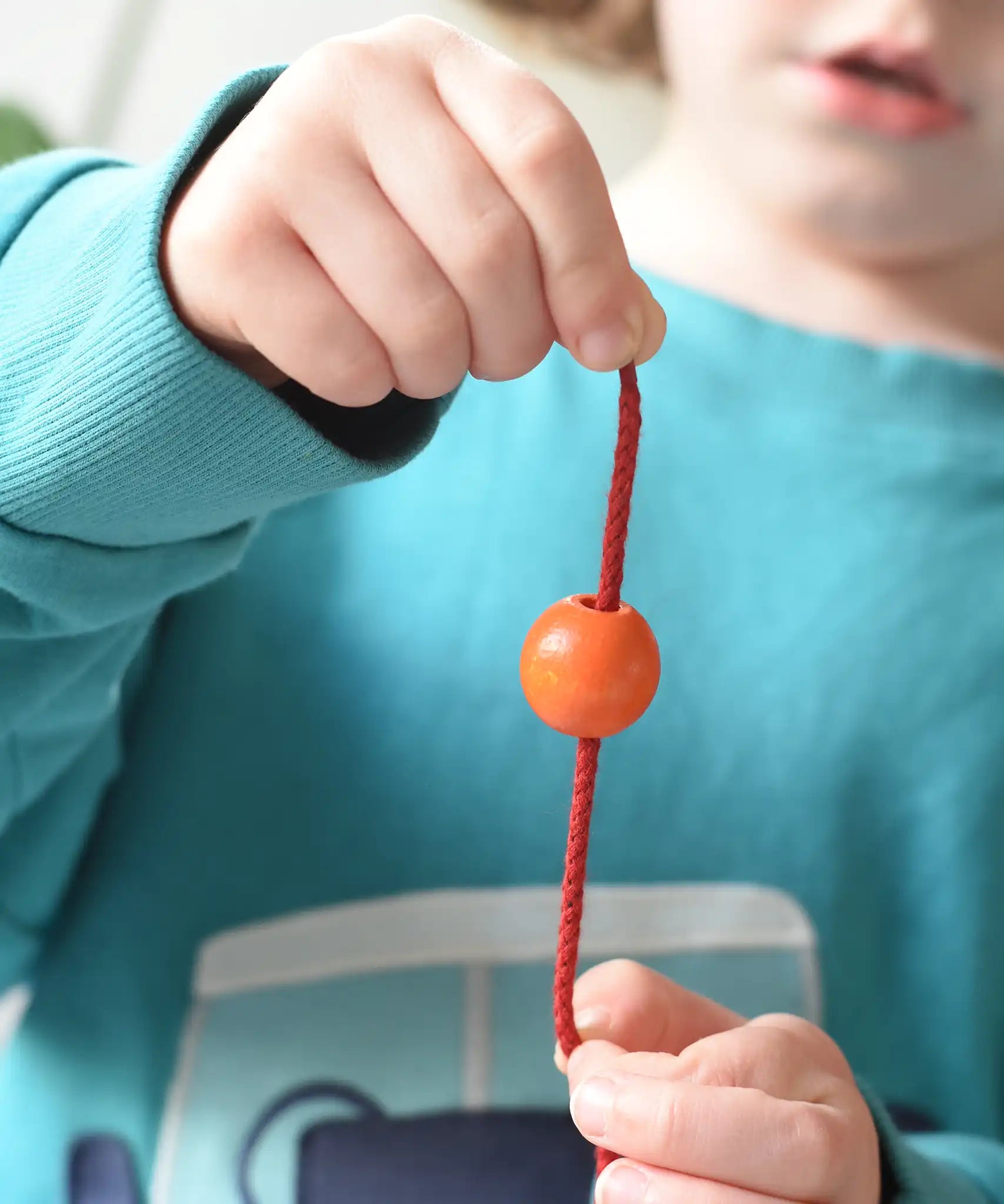 Close up of an orange bead threaded onto a red lace from the Bajo Mathematical Threading Beads Game in a child's hand.  