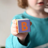 A block showing teh letter B on from the Bajo Me & You Alphabet Blocks set in a child's hand. 