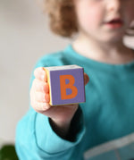 A block showing teh letter B on from the Bajo Me & You Alphabet Blocks set in a child's hand. 