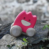 A pink coloured Bajo wooden ghost toy placed on a stone wall outdoors.  The toy has a pink painted body and natural wooden wheels. These heirloom quality toys by Bajo are part of a wide range of wooden toys available here at Babipur. 