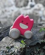A pink coloured Bajo wooden ghost toy placed on a stone wall outdoors.  The toy has a pink painted body and natural wooden wheels. These heirloom quality toys by Bajo are part of a wide range of wooden toys available here at Babipur. 