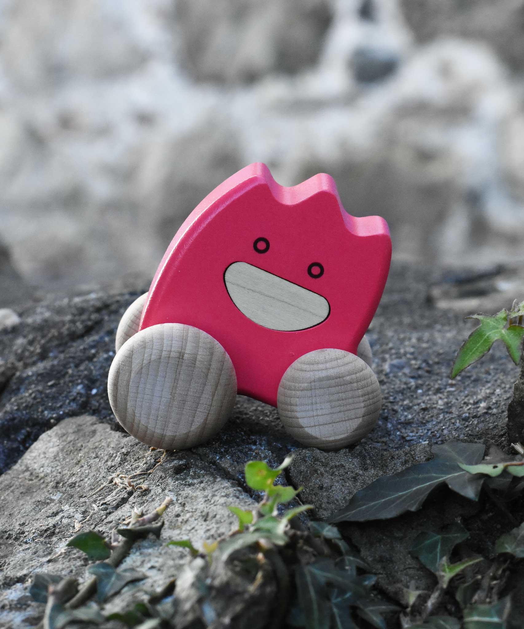 A pink coloured Bajo wooden ghost toy placed on a stone wall outdoors.  The toy has a pink painted body and natural wooden wheels. These heirloom quality toys by Bajo are part of a wide range of wooden toys available here at Babipur. 