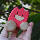 A pink coloured Bajo wooden ghost in an adult's hand.  The toy has a pink painted body and natural wooden wheels. These heirloom quality toys by Bajo are part of a wide range of wooden toys available here at Babipur. 
