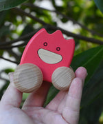 A pink coloured Bajo wooden ghost in an adult's hand.  The toy has a pink painted body and natural wooden wheels. These heirloom quality toys by Bajo are part of a wide range of wooden toys available here at Babipur. 
