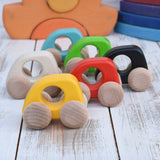 A group of Bajo handmade wooden mini cars available at Babipur. Photo is in a playroom setting with Grimms blocks in the background. Cars have unpainted wheels and cut out holes for a easy hold