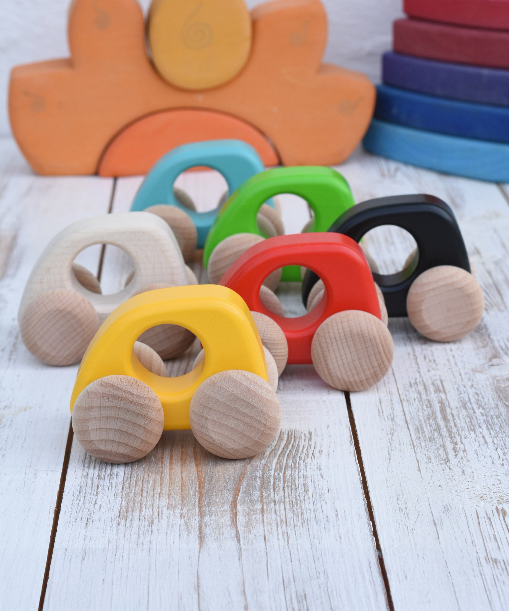 A group of Bajo handmade wooden mini cars available at Babipur. Photo is in a playroom setting with Grimms blocks in the background. Cars have unpainted wheels and cut out holes for a easy hold