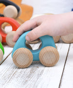A child's hand holding the Bajo hand made wooden light blue mini style car in a playroom setting showing scale. 