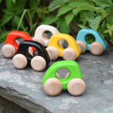 A group of Bajo handmade wooden mini cars available at Babipur. Photo is in a outdoor setting with greenery in a background. Cars have unpainted wheels and cut out holes for a easy hold