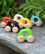 A group of Bajo handmade wooden mini cars available at Babipur. Photo is in a outdoor setting with greenery in a background. Cars have unpainted wheels and cut out holes for a easy hold