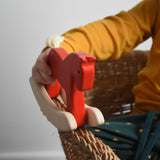 A close up of a child's hand holding a red coloured Bajo Mini Rocking Horse. The child is sitting in a wicker chair. These heirloom quality toys by Bajo are part of a wide range of wooden toys available here at Babipur. 